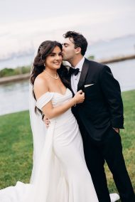 Bride in off-shoulder white gown and pearl necklace embraces groom in black tuxedo as he kisses her cheek near waterfront in New York, NY.