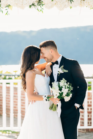 Bride and groom in elegant wedding attire touching foreheads under a white floral canopy with a waterfront and mountain backdrop in New York, NY.