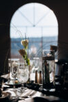 Wedding table with floating candle in tall glass, white roses, crystal glassware, and silverware in front of an arched window with daylight.