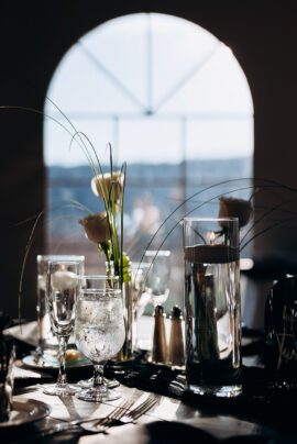 Wedding table with floating candle in tall glass, white roses, crystal glassware, and silverware in front of an arched window with daylight.