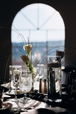 Wedding table with floating candle in tall glass, white roses, crystal glassware, and silverware in front of an arched window with daylight.