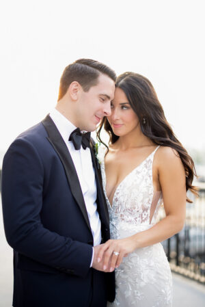 Bride in lace wedding dress and groom in black tuxedo sharing an intimate moment, holding hands on a balcony in New York, NY.