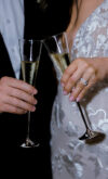 Close-up of bride and groom toasting with champagne flutes, bride wearing an embroidered lace wedding dress and diamond ring, groom in black tuxedo.