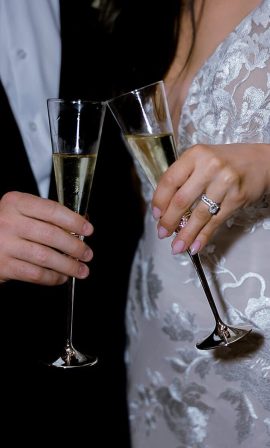 Close-up of bride and groom toasting with champagne flutes, bride wearing an embroidered lace wedding dress and diamond ring, groom in black tuxedo.