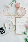 Flat lay of wedding invitation and RSVP card with gold foil edges, pearl necklace and bracelet, wedding rings on a small dish, floral earrings, rose gold perfume bottle, and pink flowers on a white textured surface.